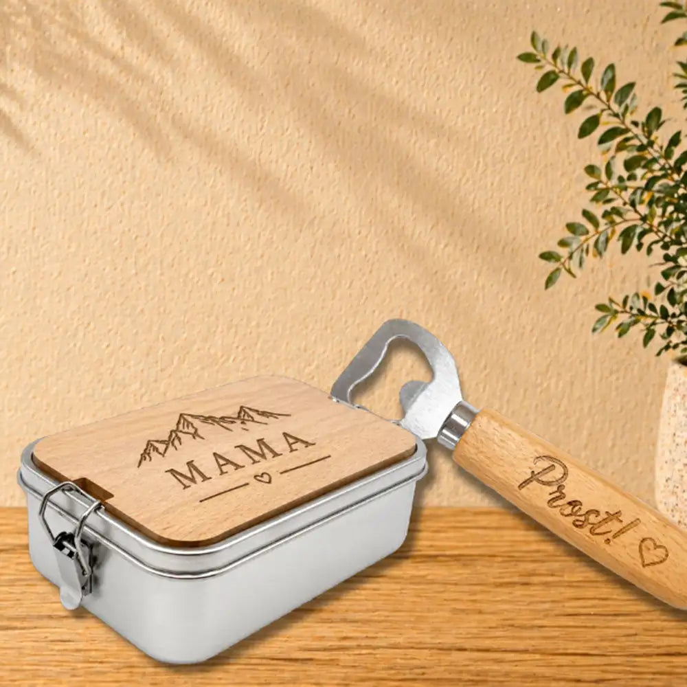 Ein Geschenkeset aus silbernem Metall und Holz mit einer rechteckigen Dose mit Holzdeckel, graviert „mama“ über Bergsilhouetten, gepaart mit einem Flaschenöffner mit natürlichem Holzgriff, beschriftet „prost!“ neben einem Herz.