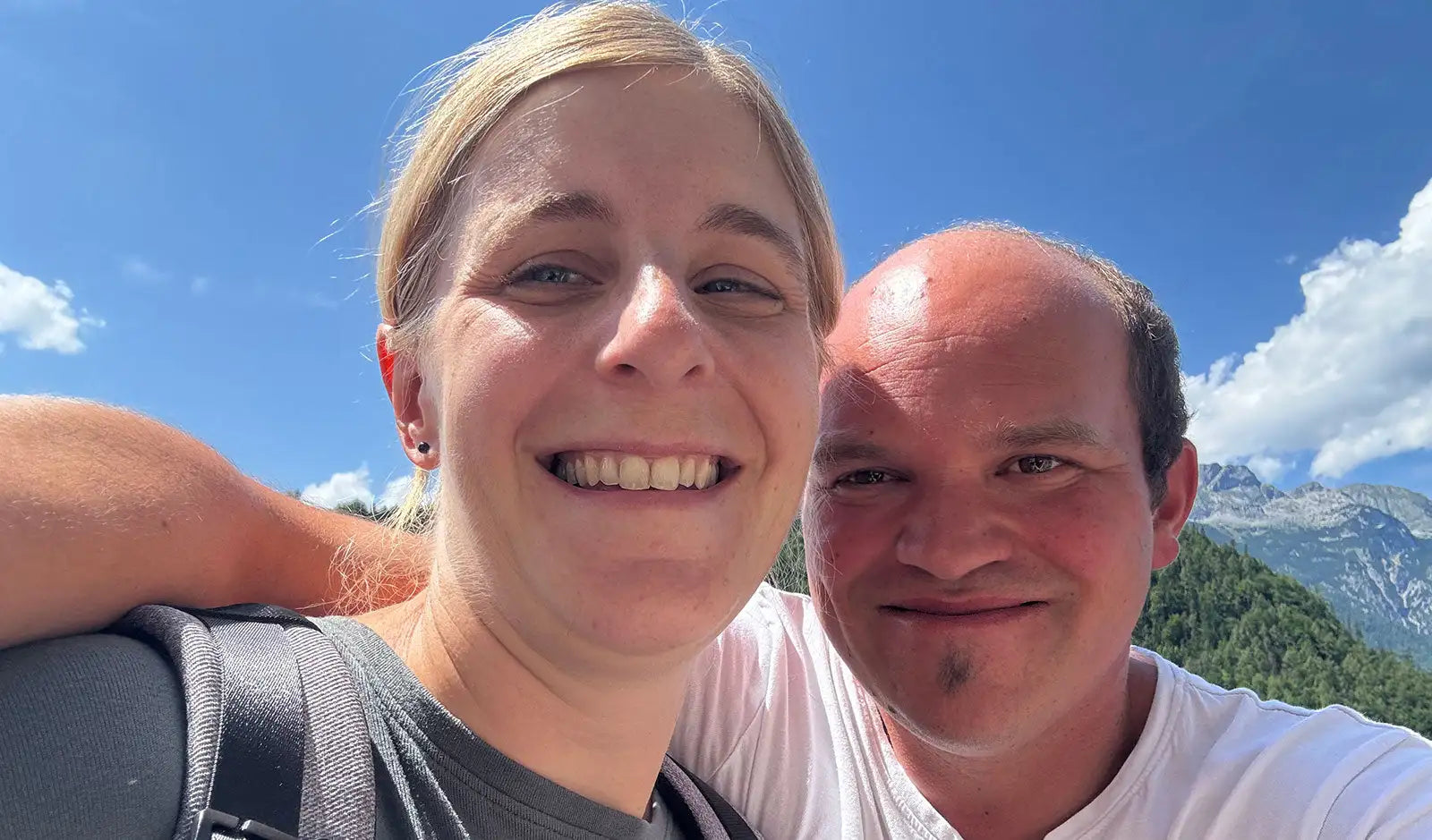 Eine Frau mit blonden Haaren und ein Mann mit kurzen dunklen Haaren posieren zusammen im Freien, beide lächeln unter einem hellblauen Himmel.